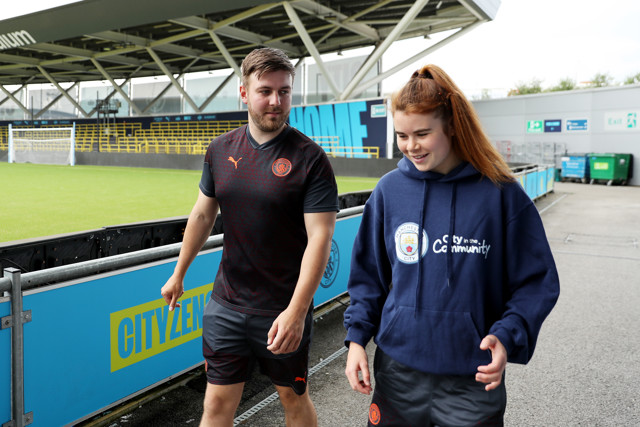 Two people, one wearing a Manchester City training kit and the other in a Manchester City Community hoodie, are walking beside the field inside a stadium.