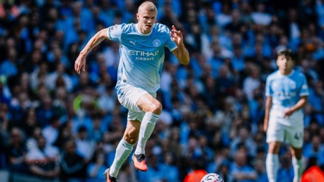 Manchester City player in action during a match, wearing the team's kit, with a crowd in the background.