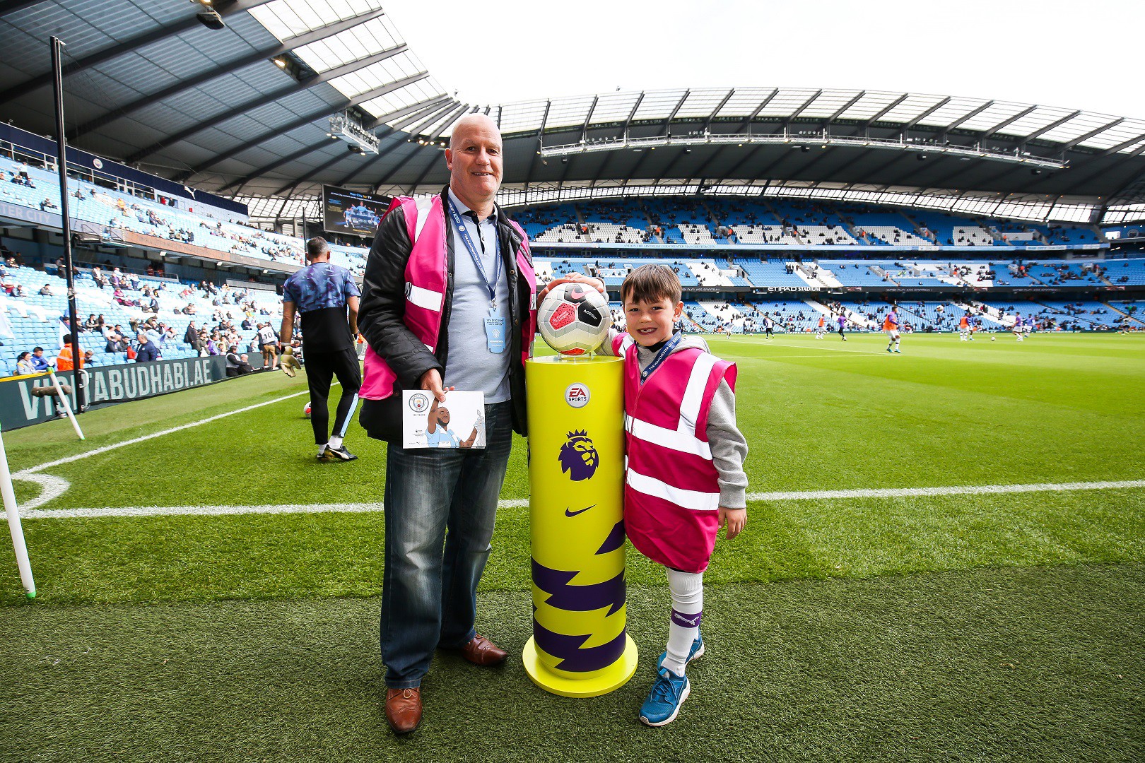 Two people, one adult and one child, standing at the Etihad Stadium near the pitch with a Premier League match ball on a pedestal. Both are wearing pink vests with lanyards.