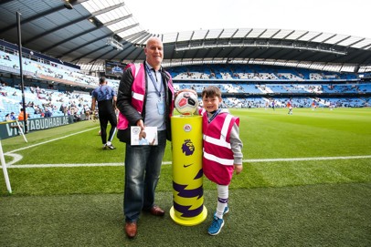 Two people, one adult and one child, standing at the Etihad Stadium near the pitch with a Premier League match ball on a pedestal. Both are wearing pink vests with lanyards.