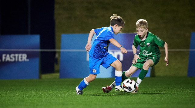 Two children in blue and green soccer kits are engaged in a football match, focusing on the ball, on a grass field.