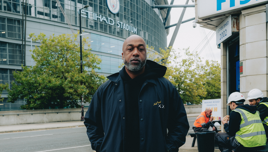 A person stands in front of Etihad Stadium wearing a black jacket, with construction workers in the background.