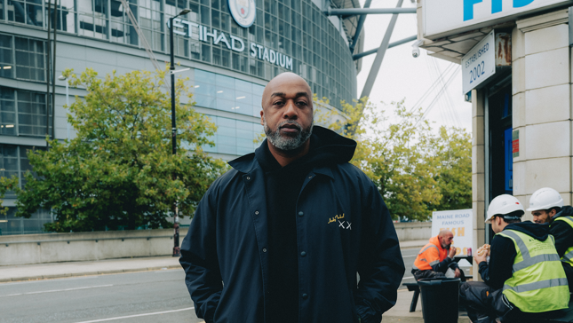 A person stands in front of Etihad Stadium wearing a black jacket, with construction workers in the background.