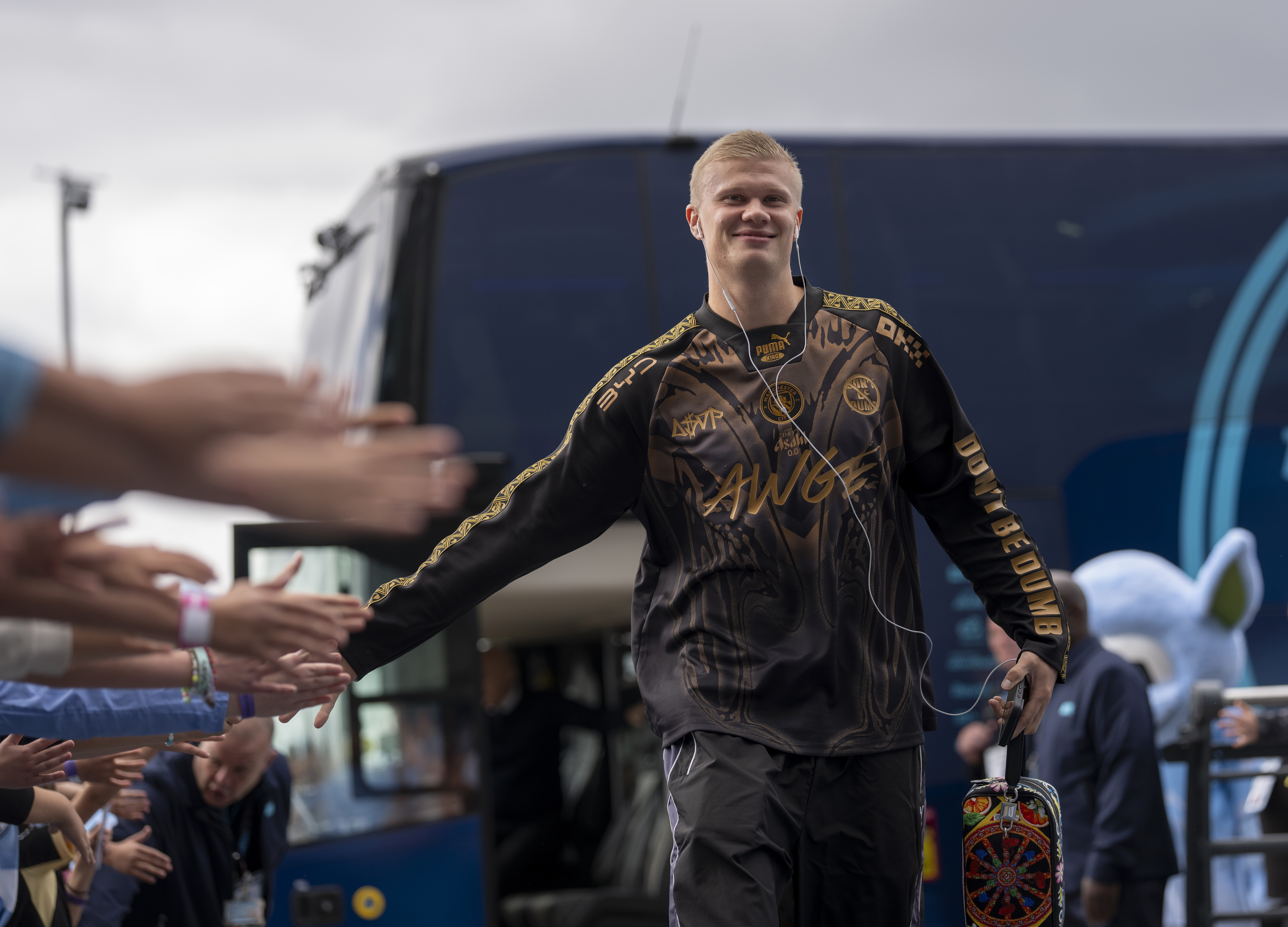 A smiling soccer player wearing headphones and casual attire, greeting a crowd with outstretched hands as he walks past a team bus.