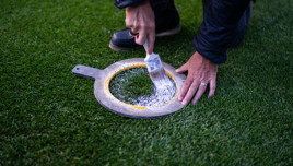 Person using a brush to apply white paint through a stencil onto grass, forming a circular mark.