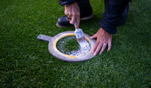 Person using a brush to apply white paint through a stencil onto grass, forming a circular mark.