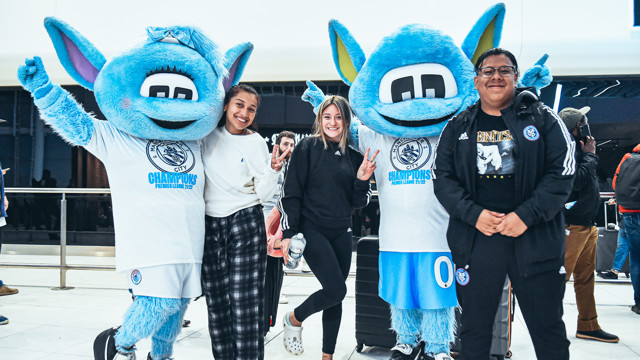 Two Manchester City mascots, Moonbeam and Moonchester, pose with fans wearing Premier League Champions 21-22 shirts.