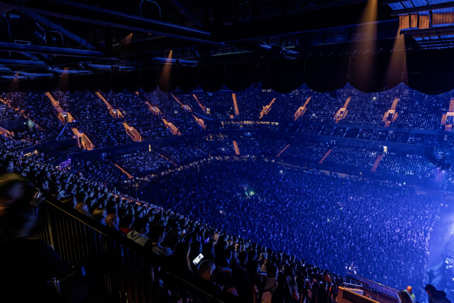 A large crowd at an indoor concert arena with blue lighting and filled seating.