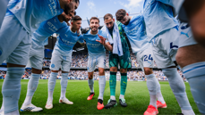 Manchester City players in a huddle on the football field wearing light blue jerseys. The stadium is filled with spectators in the background.