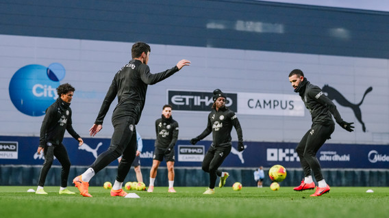 Manchester City players training at the Etihad Campus, wearing black Puma training kits. Various cones and footballs can be seen on the pitch.
