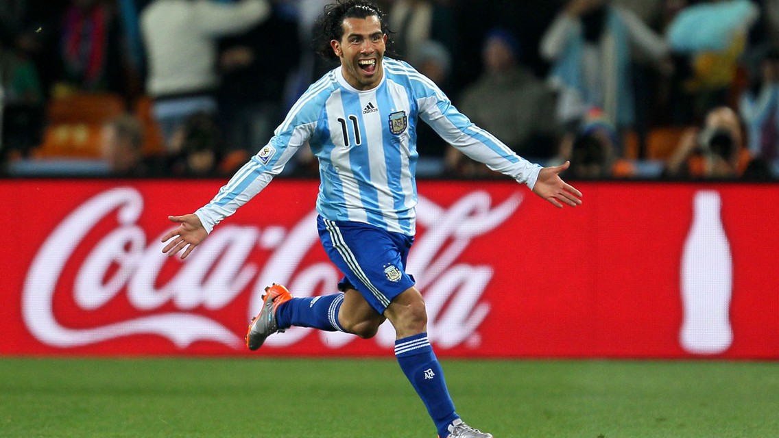 A soccer player in an Argentina jersey with the number 11 celebrates with arms spread wide on the field. A Coca-Cola advertisement is visible in the background.