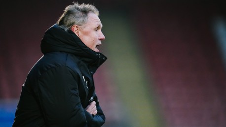 A person wearing a black jacket is standing on the sideline of a sports field, arms crossed.