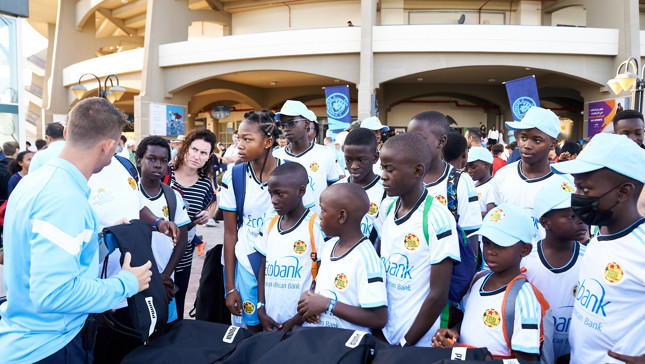 A group of children and adults gathered at a Manchester City community event, wearing white shirts and blue caps, interacting with a staff member handing out bags.