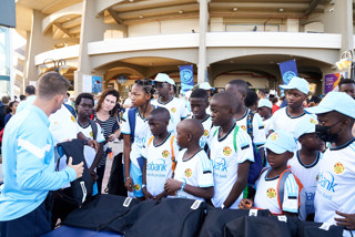 A group of children and adults gathered at a Manchester City community event, wearing white shirts and blue caps, interacting with a staff member handing out bags.