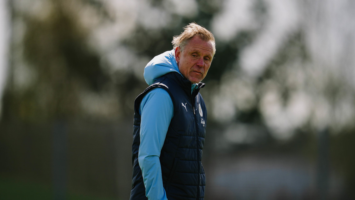 A person in a puffer vest and hoodie is standing outdoors, possibly on a sports training field. The clothing has a logo similar to Leicester City FC.