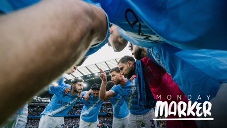 A team huddle of Manchester City players wearing light blue jerseys, captured from a low angle at the Etihad Stadium with a blurred background crowd and sky.