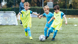 Three young children wearing soccer uniforms and training bibs are playing soccer on a field. They are running towards a soccer ball while a coach stands in the background observing them.