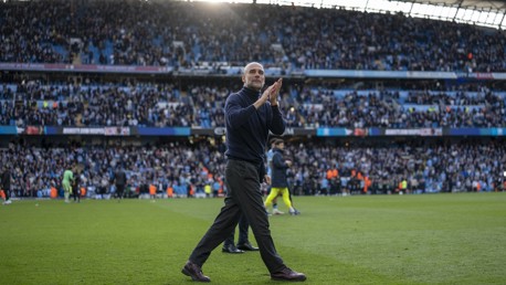 A man in a dark outfit clapping at the edge of a football field, with a crowded stadium in the background.