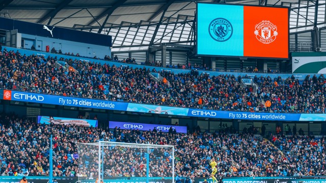 A crowded Etihad Stadium with Manchester City and Manchester United logos displayed on a scoreboard.