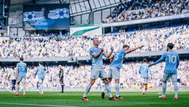 Manchester City players celebrating a goal on a football pitch with a scoreboard showing a player's image.
