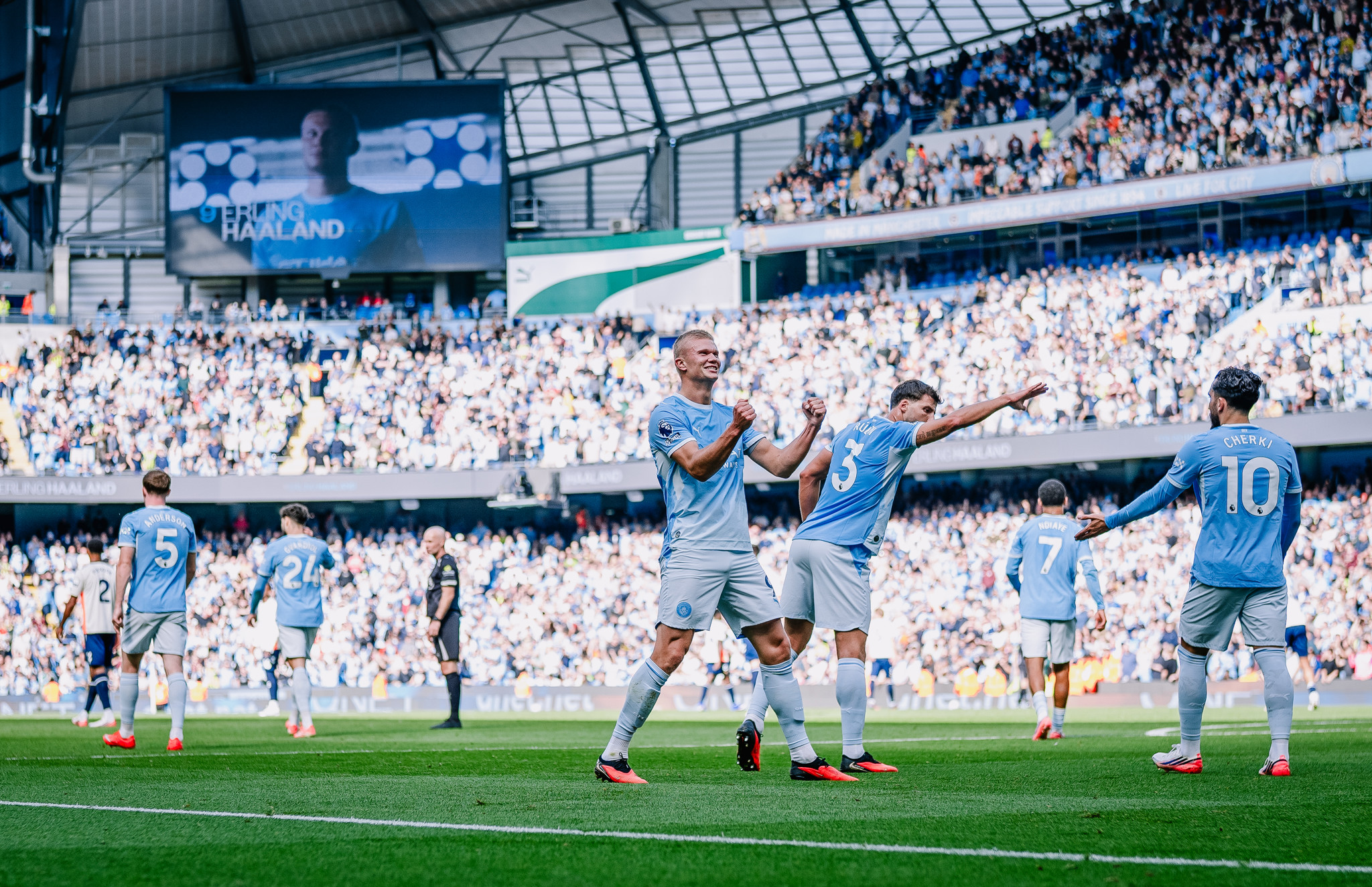 Manchester City players celebrating a goal on a football pitch with a scoreboard showing a player's image.
