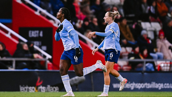 Two soccer players in light blue jerseys celebrate during a match. The player on the left appears to be holding their shirt, while the player on the right is smiling. The surrounding stadium is filled with fans.