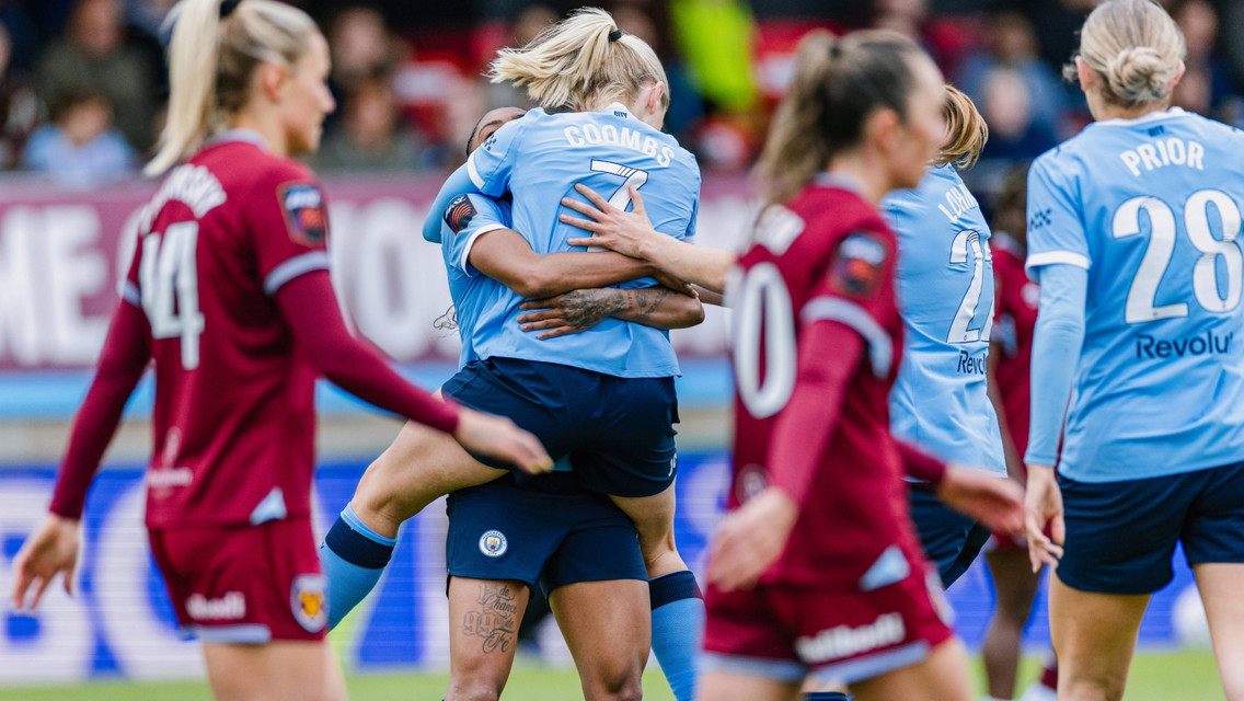 Football players celebrating a goal on the field. A player in a sky blue jersey with 'Coombs' on the back is being hugged by a teammate.