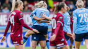 Football players celebrating a goal on the field. A player in a sky blue jersey with 'Coombs' on the back is being hugged by a teammate.