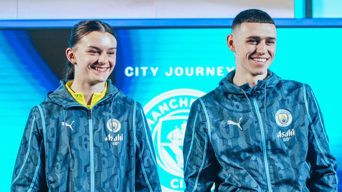 Two Manchester City players standing in front of a digital backdrop wearing Manchester City Puma branded training jackets with logos visible.