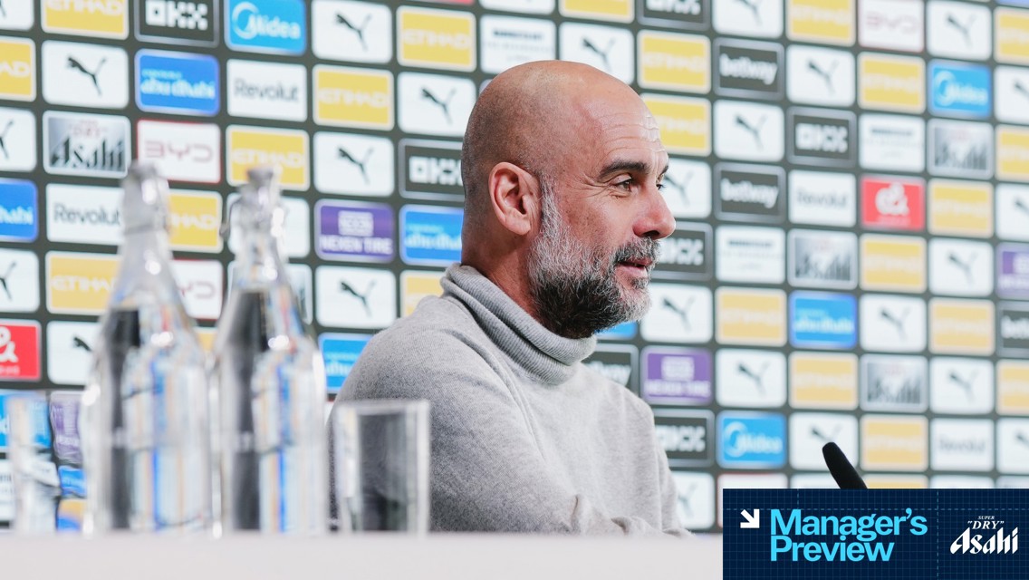 A person with blurred face speaking at a press conference with Manchester City logos displayed behind.