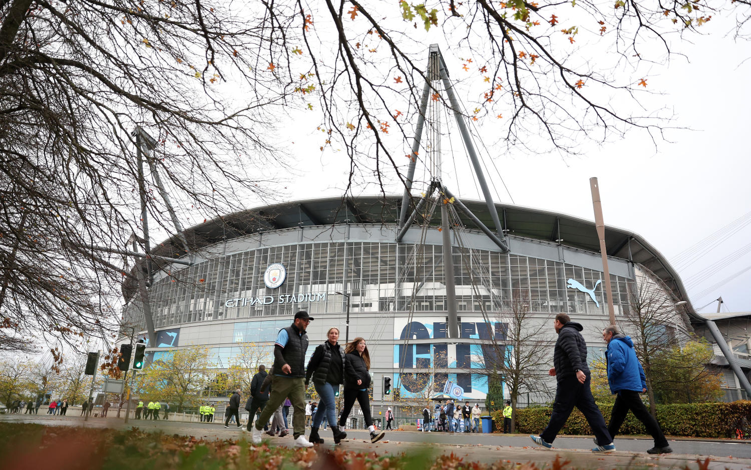 People walking near the Etihad Stadium, home of Manchester City, with trees in the foreground.