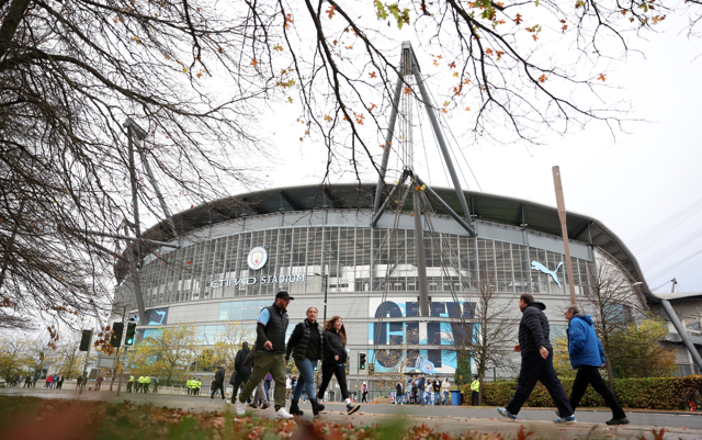 People walking near the Etihad Stadium, home of Manchester City, with trees in the foreground.
