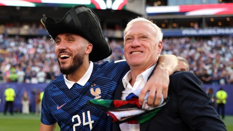 Two people celebrating wearing blue jerseys with a French rooster emblem. One of them has a number 24 jersey and a tricorn hat, while the other is holding a colorful scarf. They appear to be in a stadium.