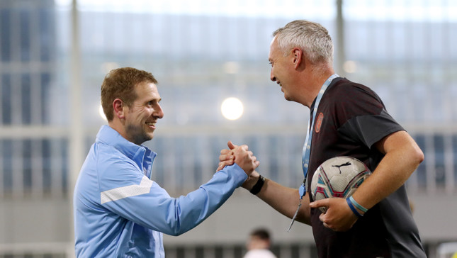 Two individuals shaking hands on a sports field, one holding a soccer ball and wearing a lanyard.