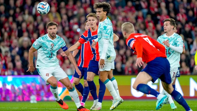 Football match between teams wearing Portugal and Norway jerseys. The ball is in the air with players focused on it during a game, captured in a stadium with a cheering crowd in the background.