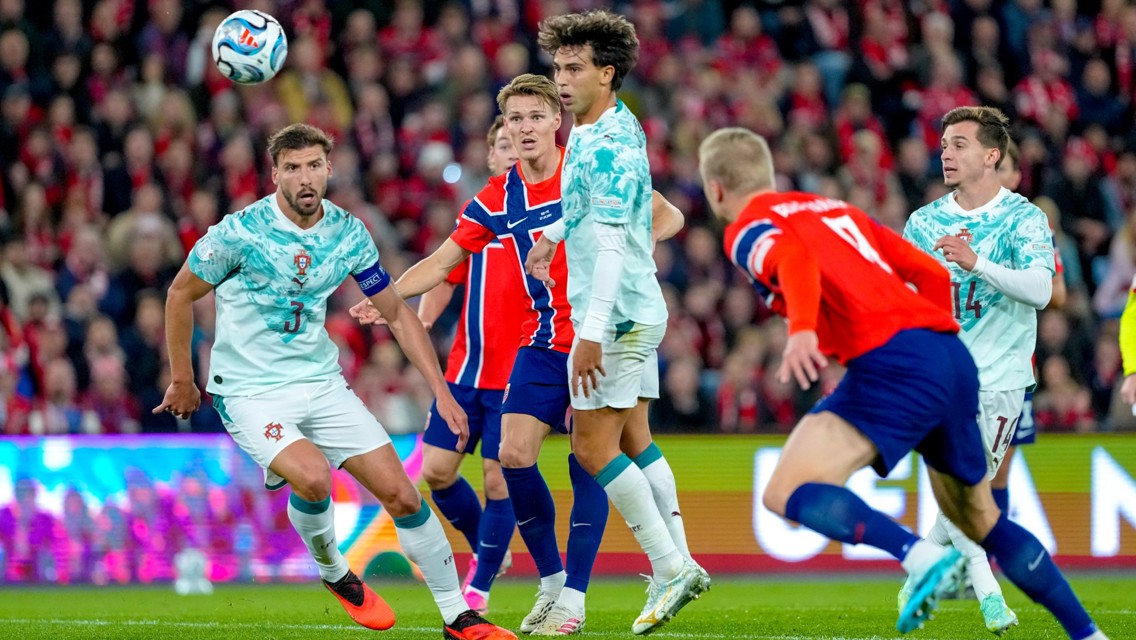 Football match between teams wearing Portugal and Norway jerseys. The ball is in the air with players focused on it during a game, captured in a stadium with a cheering crowd in the background.
