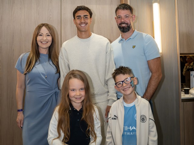 A group of five people, including three adults and two children, are posing together indoors. One child is wearing glasses and has a painted face. The adults are dressed in blue and gray attire. A Manchester City logo is visible on one child's jacket.
