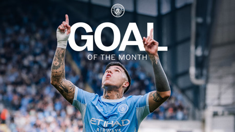 A footballer in a Manchester City jersey celebrates with hands pointing upwards after scoring the Goal of the Month.