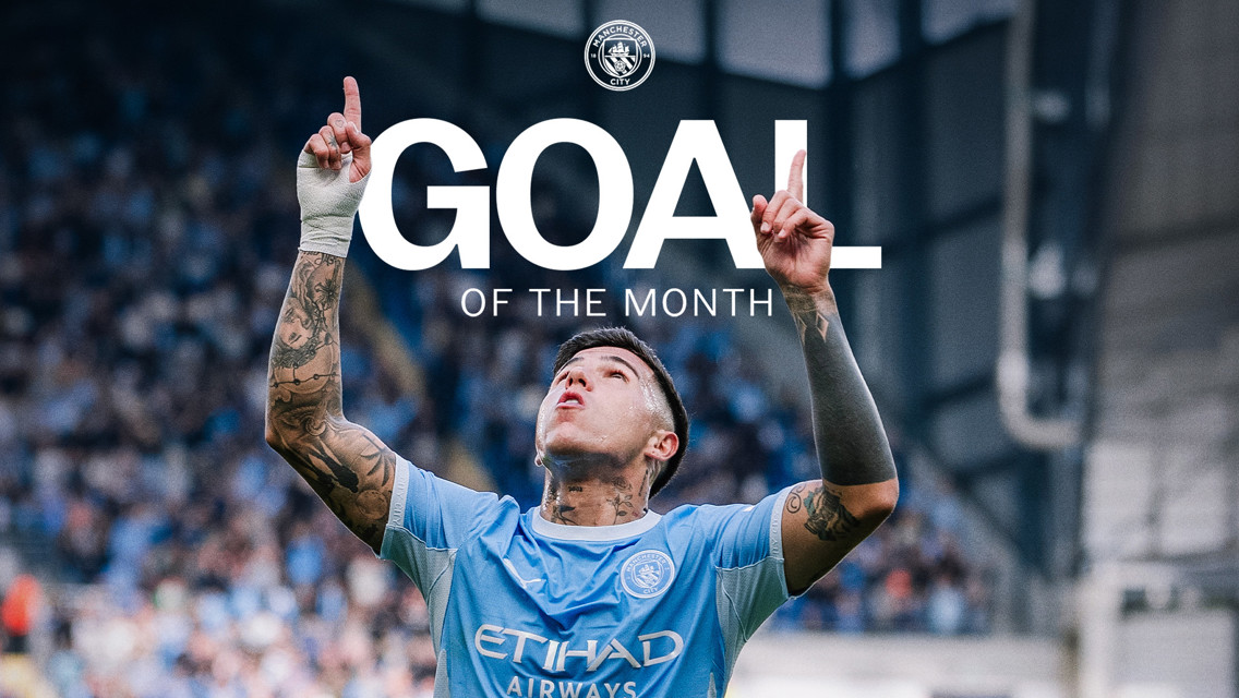 A footballer in a Manchester City jersey celebrates with hands pointing upwards after scoring the Goal of the Month.