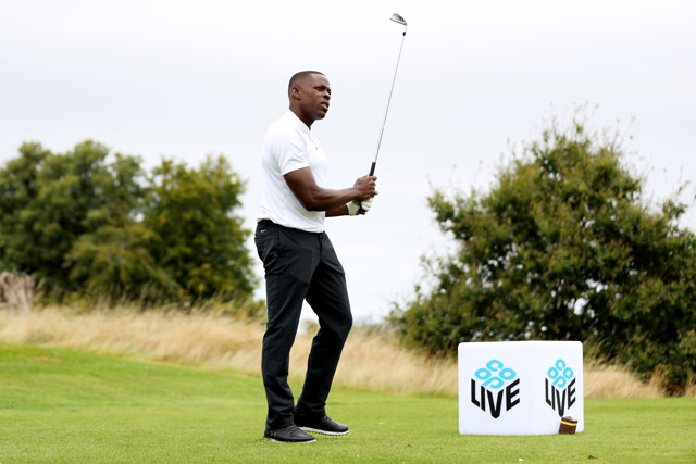 A golfer wearing a white shirt and black pants is seen preparing to swing on a golf course. There is a LIV tournament logo cube marker next to the golfer. The background includes greenery and a cloudy sky.