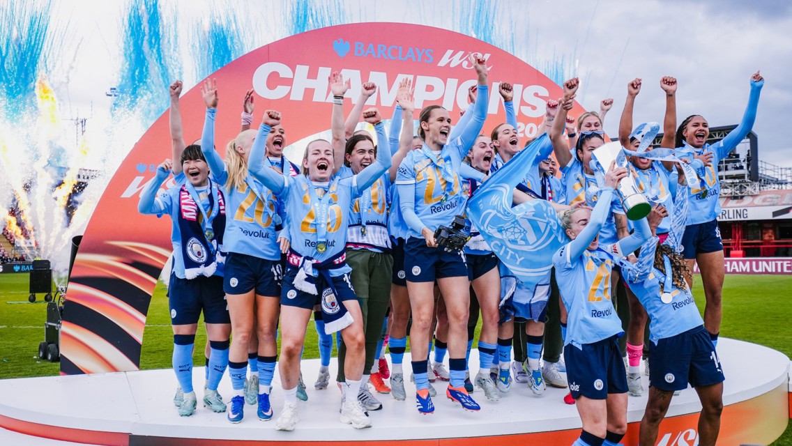 A women's football team celebrating a championship victory with fireworks in the background, wearing blue jerseys and holding a trophy.