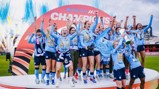 A women's football team celebrating a championship victory with fireworks in the background, wearing blue jerseys and holding a trophy.