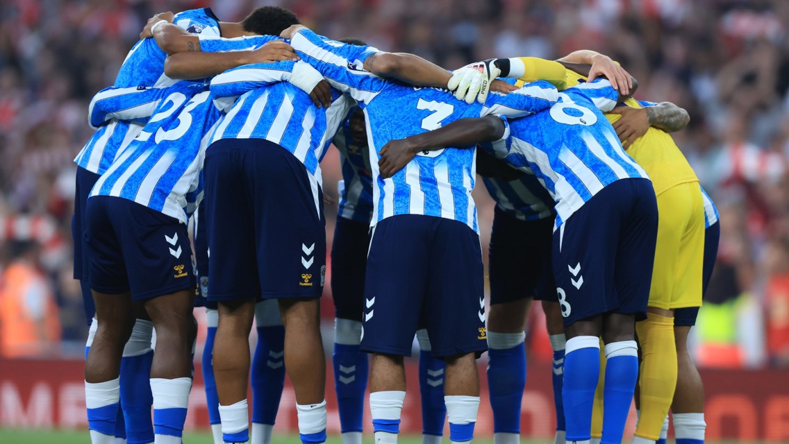 A soccer team in blue and white striped jerseys huddles together on the field before a match.