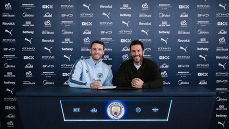 Two individuals at a contract signing table with Manchester City branding in the background.