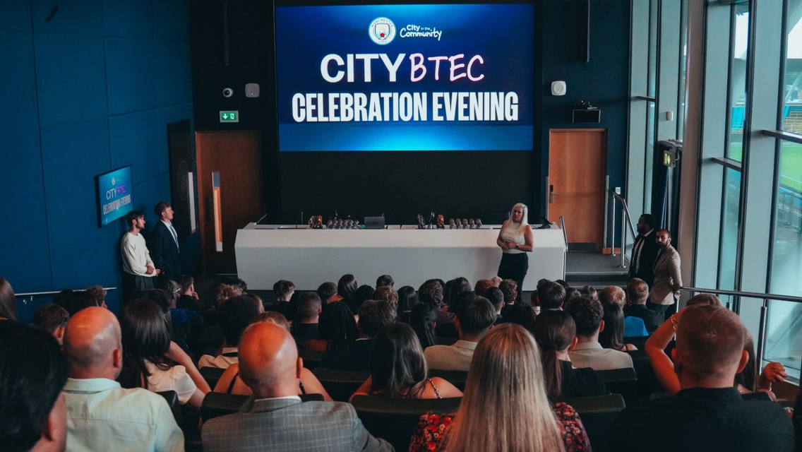 A group of people attending a City BTEC Celebration Evening hosted by City in the Community. Trophies are displayed on a table in front of a large screen showing the event title.