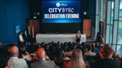 A group of people attending a City BTEC Celebration Evening hosted by City in the Community. Trophies are displayed on a table in front of a large screen showing the event title.