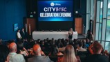 A group of people attending a City BTEC Celebration Evening hosted by City in the Community. Trophies are displayed on a table in front of a large screen showing the event title.