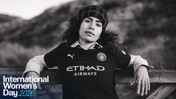 A woman wearing a Manchester City football jersey with a blurred face, leaning against a wooden rail. The image features an International Women's Day 2026 graphic.