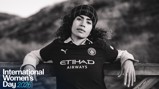 A woman wearing a Manchester City football jersey with a blurred face, leaning against a wooden rail. The image features an International Women's Day 2026 graphic.