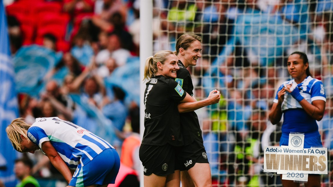 Two players in black jerseys celebrate while one in blue and white kneels. Sign in lower right reads 'Adobe Women's FA Cup Winners 2026'.